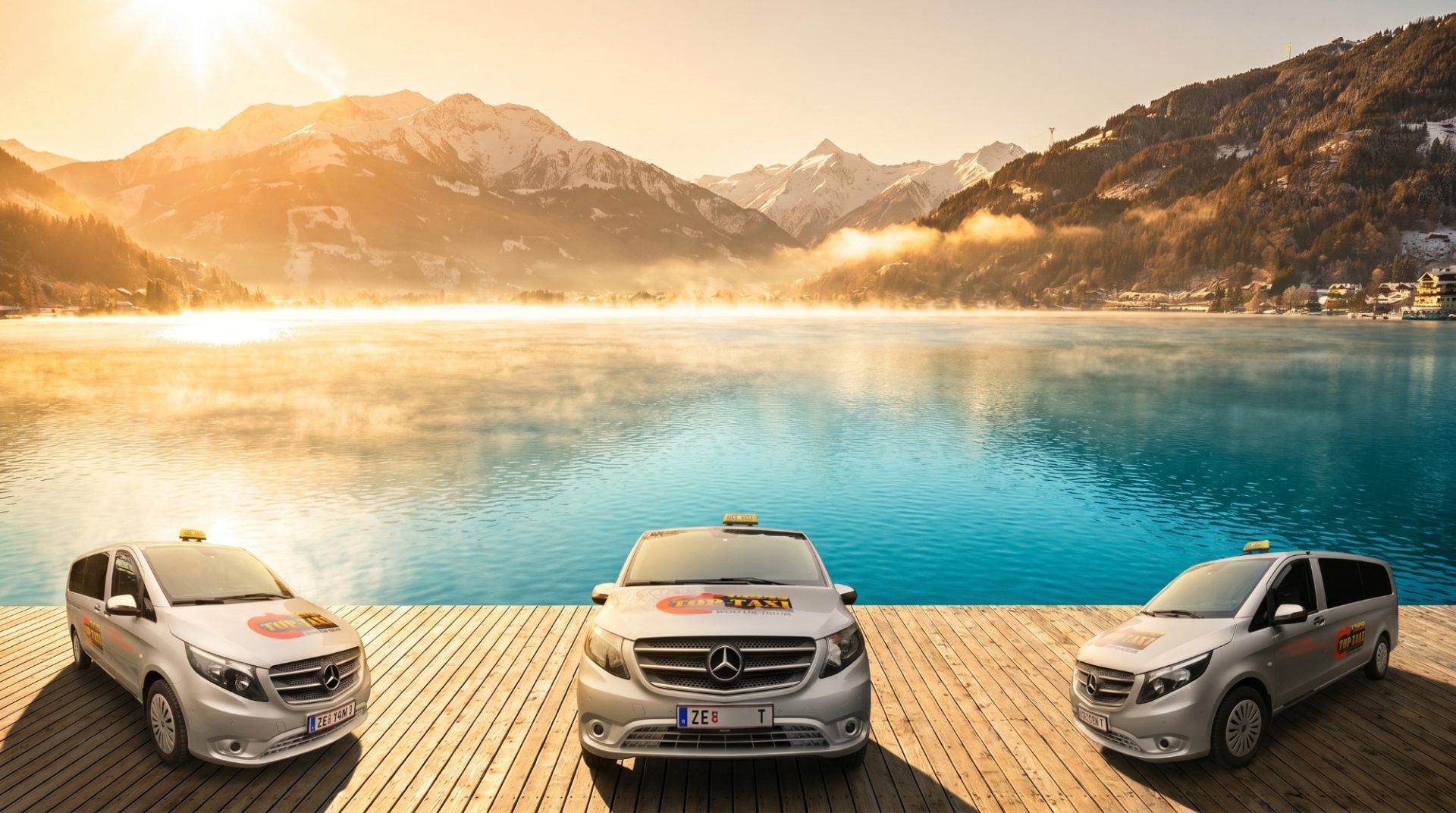 Three TOP TAXI Mercedes vans on the wooden lake pier at sunrise in Zell am See, with snow-covered mountains behind